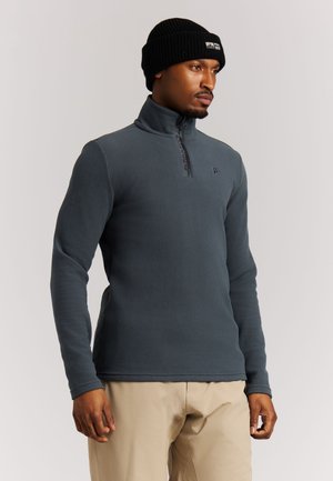 Hombre con un forro polar gris oscuro con media cremallera, pantalones beige y un gorro de punto negro, mirando ligeramente hacia la derecha.