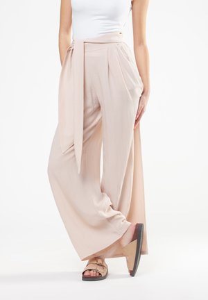 Donna con pantaloni beige a vita alta e gamba larga, con cintura in tessuto abbinata, top bianco senza maniche e sandali beige.