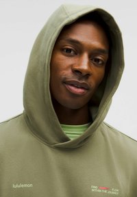 Sweatshirt olive avec cordon de serrage et poche avant, arborant le logo lululemon et un texte, superposé sur une chemise vert clair.
