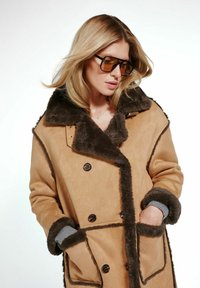 Beżowy zamszowy płaszcz z brązowym sztucznym futrem przy kołnierzu i wykończeniach, zapinany na podwójne guziki, z kieszeniami z przodu. Widoczne szare dzianinowe mankiety.