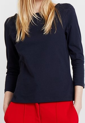 Femme portant un haut noir à manches longues avec des épaules bouffantes et un pantalon rouge vif, debout les mains dans les poches devant un fond uni.