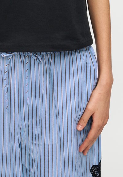 Χέρι ακουμπισμένο δίπλα σε ανοιχτό μπλε ριγέ παντελόνι με κορδόνι στη μέση και μαύρη λεπτομέρεια δαντέλας, φορώντας μαύρο πουκάμισο.