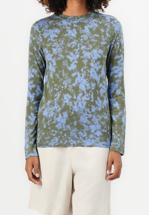 Osoba ubrana w długorękawową koszulkę w zielono-niebieskie wzory tie-dye w połączeniu z luźnymi, jasnobeżowymi szortami na białym tle.