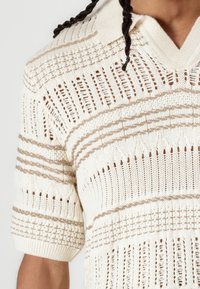 Polo en tricot crème avec des rayures horizontales beiges, présentant un motif en open weave, des manches courtes et un col texturé.