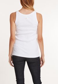 Witte mouwloze tanktop met een geribbelde textuur en een ronde halslijn, gedragen met donkerblauwe jeans, met een klein label aan de achterkant.