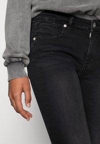 Vaqueros negros de corte ajustado, con un diseño clásico de cinco bolsillos, herrajes plateados y una textura de denim suave. Acentuados por una costura sutil.