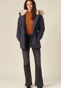 Chaqueta acolchada de color marino con capucha forrada de piel sintética, diseño acolchado y cierres a presión, llevada sobre una blusa marrón de canalé y jeans oscuros de campana.