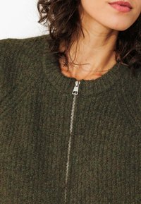 Cardigan en maille vert avec un col rond, une fermeture éclair argentée et un tissu texturé. La vue rapprochée met en évidence les détails de couture complexes.