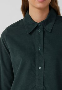 Camicia in corduroy verde scuro con colletto classico, chiusura frontale con bottoni e tessuto testurizzato. I bottoni presentano un accento di colore in contrasto.