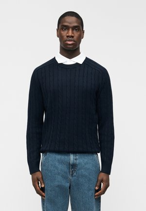 Giovane uomo di colore che indossa un maglione blu navy a trecce sopra una camicia bianca con colletto e jeans azzurri, in piedi davanti a uno sfondo bianco uniforme.