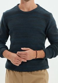 Pull en maille bleu foncé avec un col ras du cou côtelé et des manches longues, présentant un motif à rayures texturé. Bracelet marron visible au poignet.