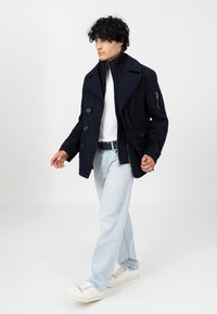 Granatowa peacoat z dużymi guzikami, nałożona na ciemny golf i białą koszulkę. Jasnoniebieskie dżinsy i białe trampki dopełniają strój.