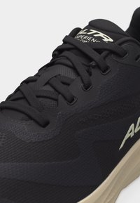 Κοντινό πλάνο μαύρου αθλητικού παπουτσιού με μαύρα κορδόνια, επάνω μέρος από δίχτυ και μπεζ σόλα, που δείχνει μέρος της επωνυμίας "ALTRA" στο πλάι.