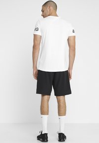 Biela športová tričko s krátkymi rukávmi a čiernym logom na ľavom ramene, v kombinácii s čiernymi šortkami a bielymi ponožkami.