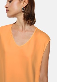 Top arancione a maniche corte e scollatura a V, realizzato in tessuto liscio. Presenta una leggera lucentezza e linee pulite, con un design minimalista. Nessun motivo visibile.