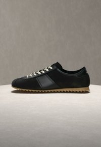 Μαύρο χαμηλό sneaker με λευκά κορδόνια, μαύρη πλαϊνή ρίγα και καφέ ανάγλυφη λαστιχένια σόλα σε ουδέτερη επιφάνεια και φόντο.