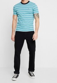 Camiseta a rayas en color verde azulado con detalles en blanco y azul marino, mangas cortas, cuello redondo clásico, combinada con pantalones negros y zapatillas negras.