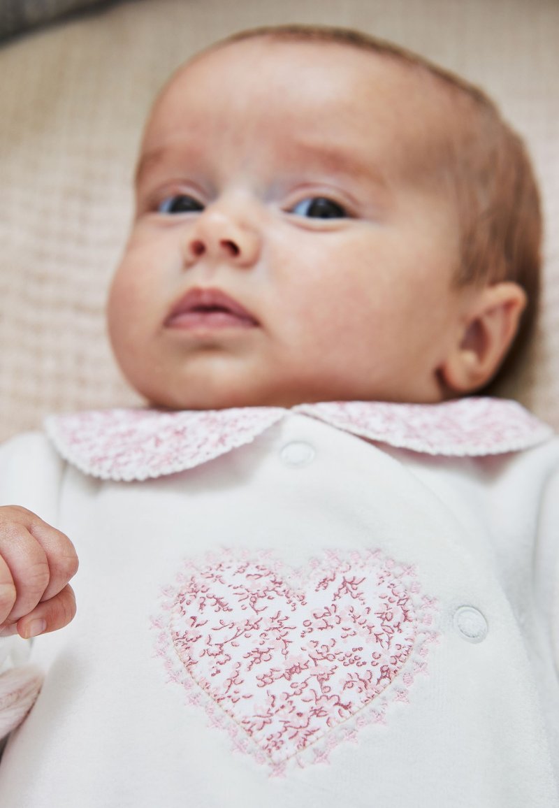 Bébé allongé portant une tenue blanche avec un cœur rose brodé et un col avec des détails en dentelle rose.
