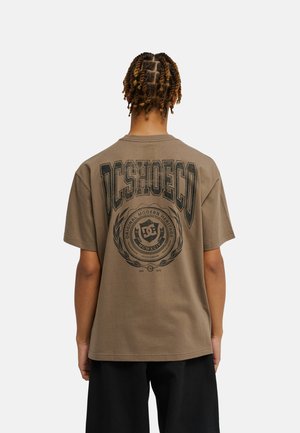 Osoba z ciasno splecionymi warkoczami odwrócona tyłem, ubrana w luźny brązowy T-shirt z dużym "DCSHOECO" i logo na plecach, połączona z czarnymi szortami.
