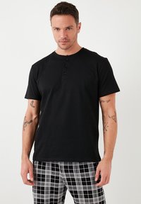 T-shirt nero a maniche corte con scollatura a girocollo e patta con tre bottoni. Tessuto di cotone morbido. Il modello indossa pantaloni a quadri bianchi e neri.