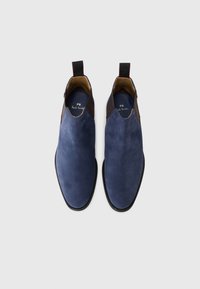 Zapatos slip-on de ante azul marino con detalles en cuero marrón, punta redonda y suela negra. Presentan una plantilla con marca y una pestaña en el talón.