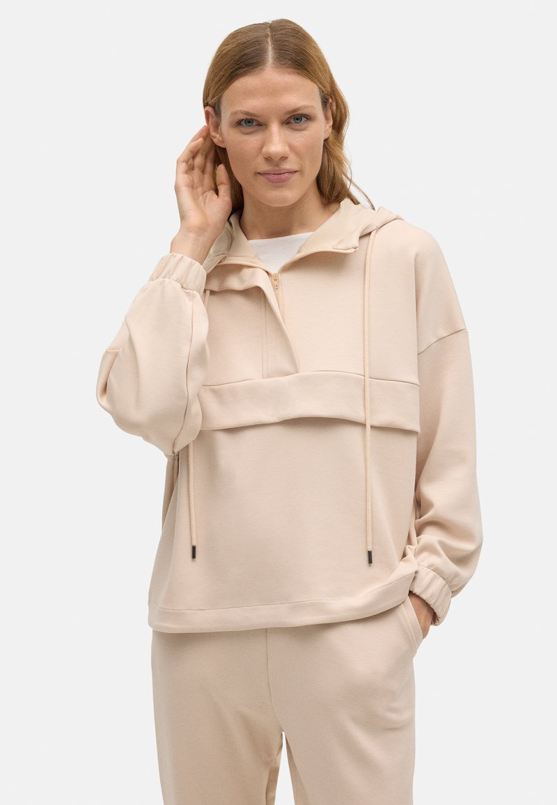 Pullover beige claro con capucha, presenta un cuello con media cremallera, bolsillo frontal y puños elásticos. Material suave con una textura lisa.