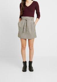 Burgundská žebrovaná halenka s dlouhým rukávem v kombinaci s béžovou kostkovanou sukní se šňůrkou v pase a bo�čními kapsami, doplněná černými kotníkovými botami.