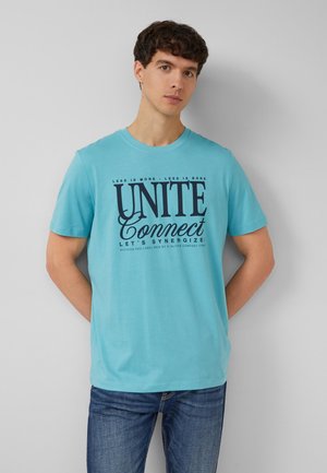Jonge man met een lichtblauw T-shirt met de tekst "UNITE Connect" en donkerblauwe jeans, staand tegen een grijze muur met de handen achter de rug.