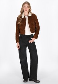 Giacca cropped in corduroy marrone con colletto in finta pelliccia crema, abbinata a jeans neri a gamba larga a vita alta e stivaletti neri.