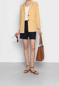 Blazer giallo chiaro con due bottoni e tasche, abbinato a pantaloni corti blu navy. Borsa a tracolla in pelle marrone e sandali color beige completano l'outfit.