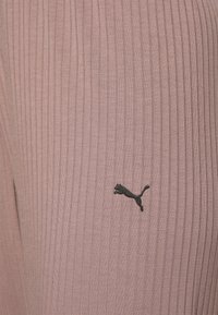 Gestreifte pinkfarbene Leggings aus weichem Stoff, mit einem schwarzen Puma-Logo auf der linken Seite. Die Textur ist glatt mit sichtbaren vertikalen Linien.
