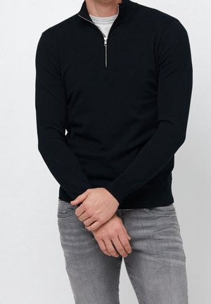 Homme portant un pull noir à manches longues avec fermeture éclair au col, par-dessus un jean gris clair, les mains légèrement jointes devant, au niveau de la taille.