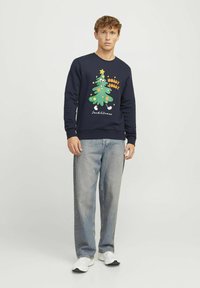 Námornícky sveter s grafickým vyobrazením vianočného stromčeka v zelenej a žltej farbe, s nápisom "HOLLY JOLLY." Kombinovaný so svetlomodrými džínsy a bielymi teniskami.