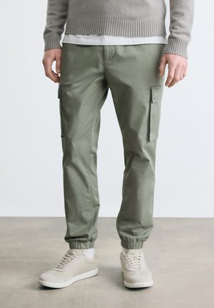 Homme portant un sweat-shirt gris clair, des baskets blanches et un pantalon cargo vert olive avec des chevilles élastiques, debout sur un sol en béton.