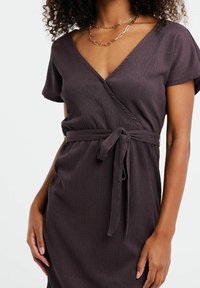 Vestido envolvente en malva oscuro con mangas cortas, escote en V y cinturón de lazo. Presenta una tela texturizada con sutiles líneas verticales.