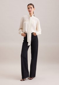 Blouse blanche avec un nœud au cou, manches longues et détails froncés, associée à un jean noir taille haute à jambes larges et des escarpins noirs.
