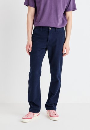 Pantalons en coton bleu marine avec une coupe droite, dotés d'une fermeture à bouton et de passants de ceinture, associés à un T-shirt violet et des baskets roses.