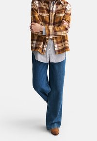 Camisa de franela a cuadros marrón y crema superpuesta sobre una camisa de botones a rayas azul y blanca, combinada con vaqueros de pierna ancha azules y zapatos marrones.