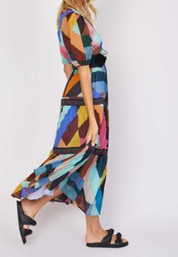 Robe longue colorée à motifs avec manches courtes bouffantes, motifs géométriques diagonaux en bleu, rose et orange, associée à des sandales slides noires.