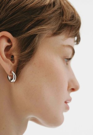 Joven con cabello corto y castaño, que lleva un pequeño pendiente de aro plateado, mostrado de perfil sobre un fondo claro.