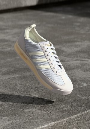 Světlé šedé a krémové tenisky Adidas s pleteným svrchním dílem a zežloutlou podrážkou, zobrazené ve vzduchu nad betonovým povrchem.