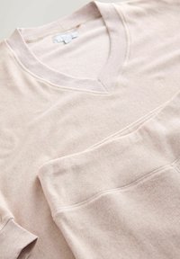 Jasno beżowy sweter z dekoltem w serek, wykonany z miękkiej, teksturowanej tkaniny. Posiada ściągacze przy mankietach oraz kieszeń naszywaną. Gładkie szwy i stonowany design.