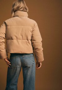 Beżowa kurtka puchowa z materiału kordowego, z wysokim kołnierzem i krótkim fasonem. Noszona na niebieskich dżinsach o klasycznym pięciopocketowym kroju.