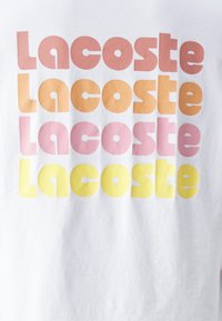 Bílý tričko s opakujícím se textem "Lacoste" v různých odstínech růžové, oranžové a žluté. Tučně zaoblená písmena vytvářejí vrstvený efekt.