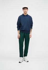 Marineblå hettegenser med krage, grønne treningsbukser, hvite joggesko og en lys caps. Enkel design med broderi foran.