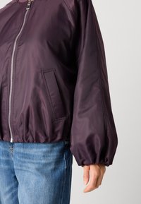Giacca bomber viola con chiusura lampo, polsini elastici e tasca laterale. Tessuto liscio, leggero e con dettaglio di orlo arricciato.