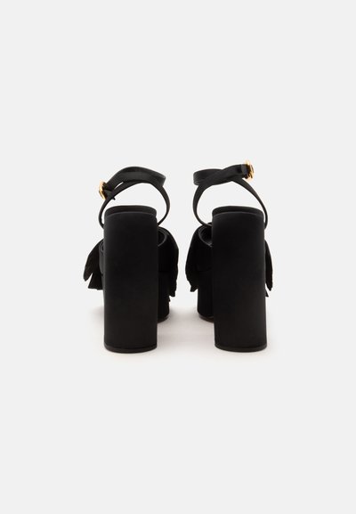 Sandales noires à talons hauts épais et brides à la cheville avec boucles dorées, vues de dos sur un fond blanc.