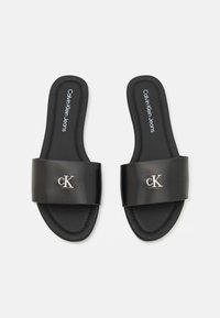 Infradito neri con una fascia larga che presenta il logo "CK" in argento. La soletta è caratterizzata da dettagli testurizzati e la suola è piatta e liscia.