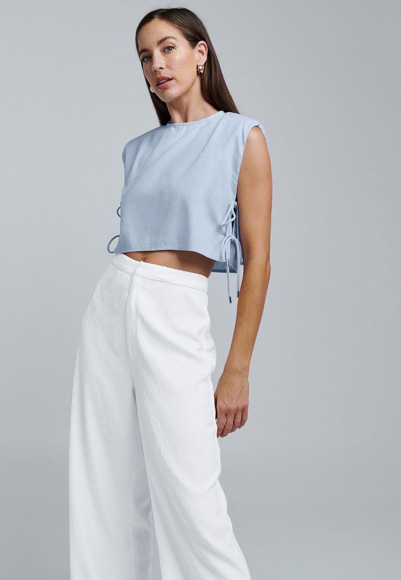 Lyseblå cropped top med ærmeløst design og sidebindinger, parret med hvide vide bukser i tekstureret stof.