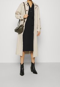 Frau in einem beigen Trenchcoat über einem schwarzen, taillierten Kleid, mit schwarzen Knöchelstiefeln und einer olivgrünen Schultertasche.